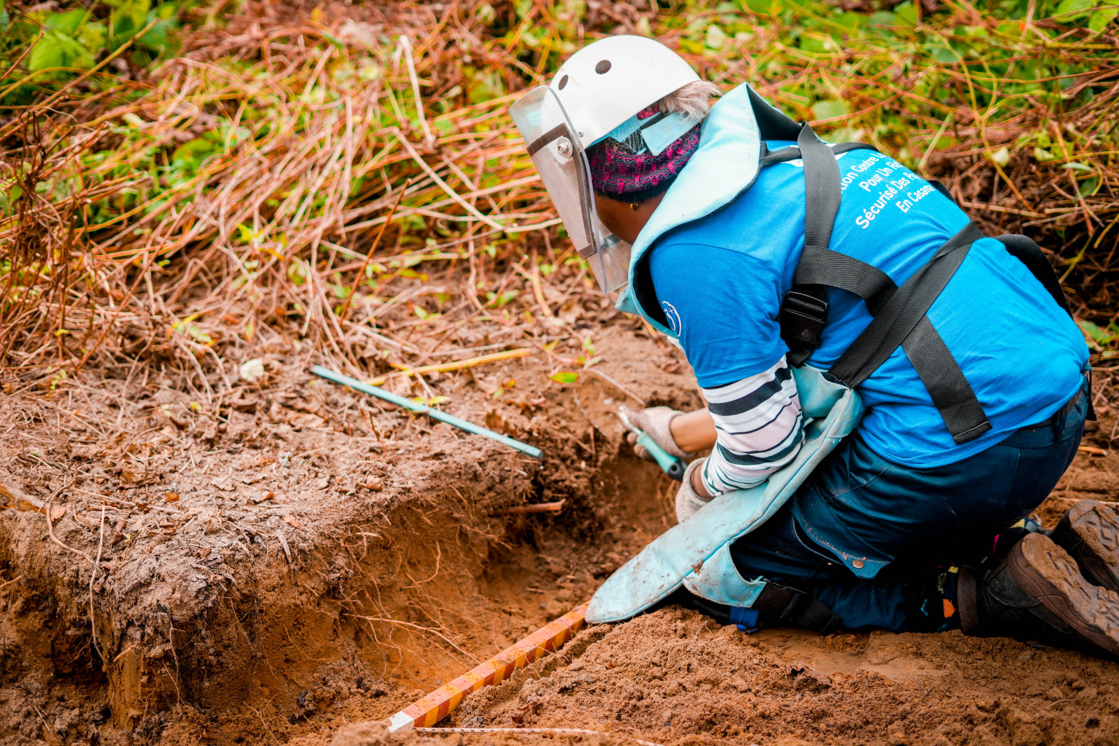 Trois démineurs et une démineuse en tenue de protection se tiennent sur un chemin de terre, entourés de végétation luxuriante.