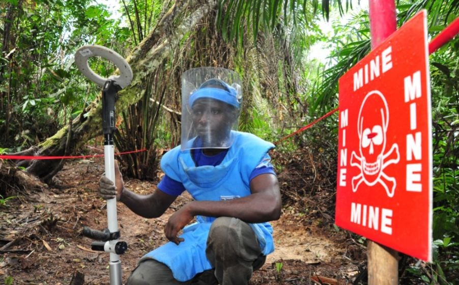 Deminer sitting with demining equipment in front of a sign that says dangerous