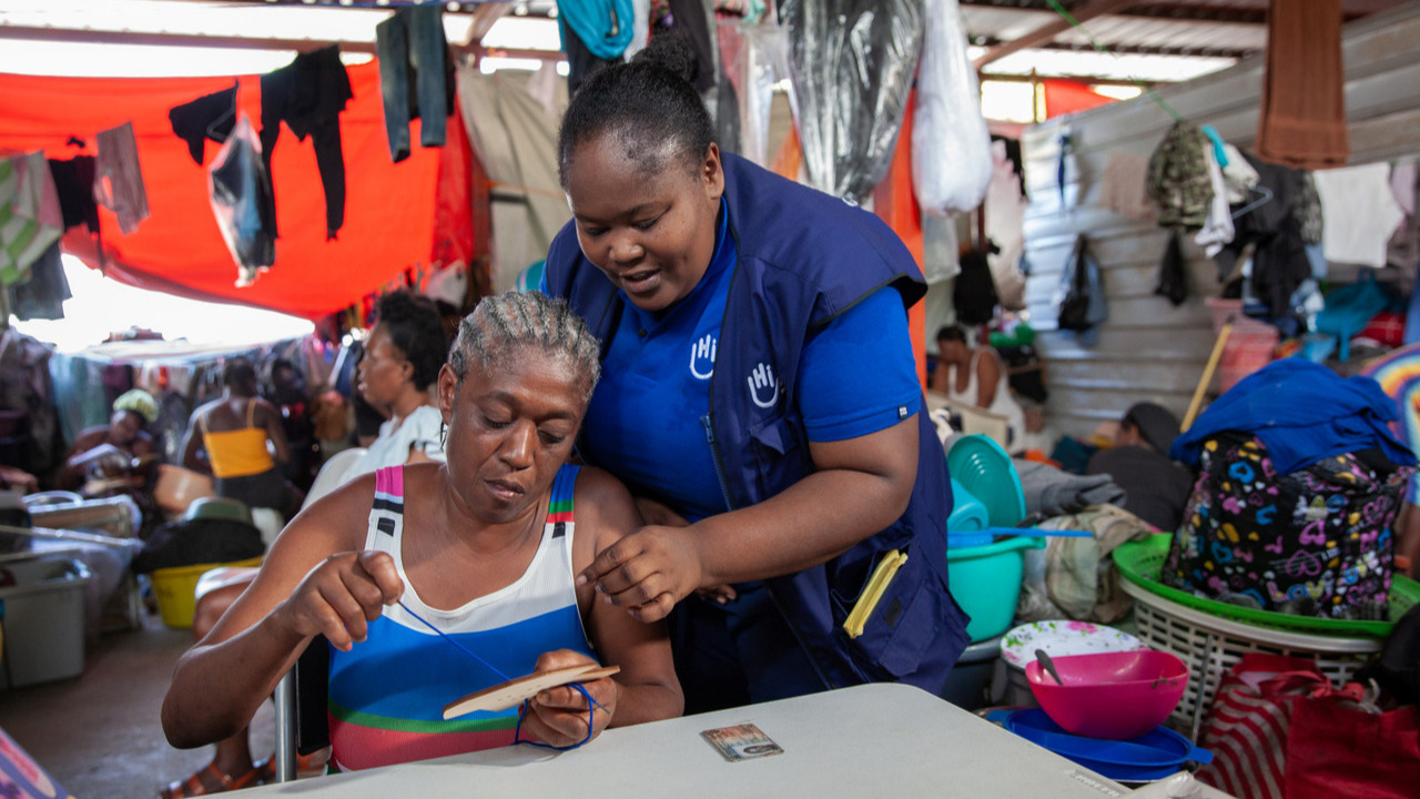A woman sitting down is threading a piece of thread through a wooden frame, guided by another woman standing next to her.