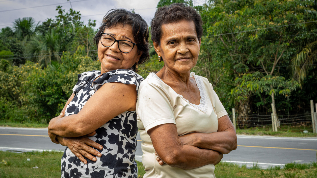 Two women are standing back-to-back, striking a confident pose, and looking at the camera with a smile.