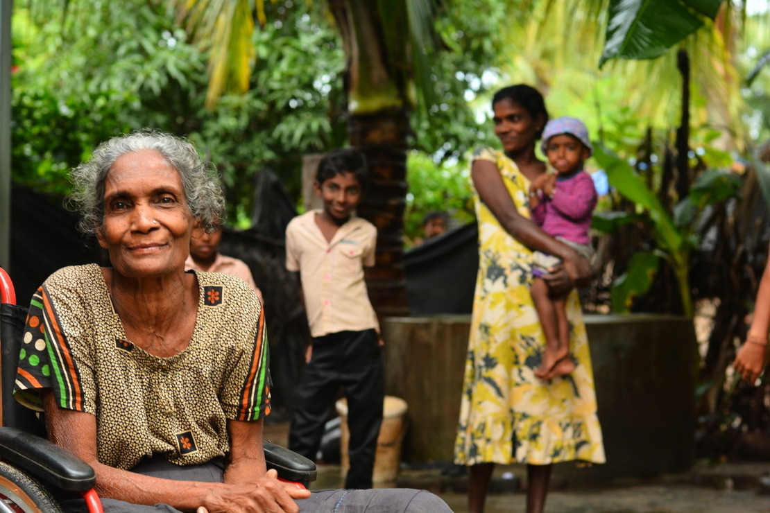 Une femme pose en fauteuil roulant au premier plan avec le reste de sa famille dont un bébé en arrière plan sous des arbre