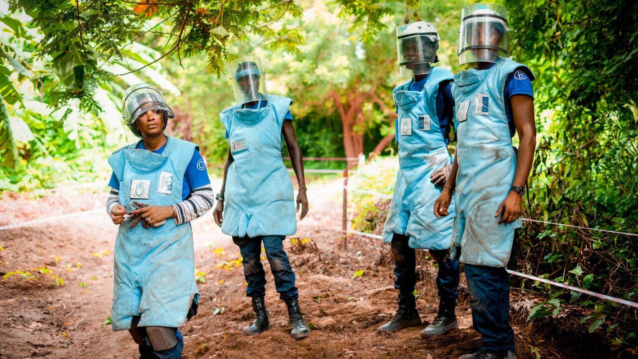 Quatre personnes habillées en tenue de protection de déminage se tiennent sur un chemin de terre ombragé.