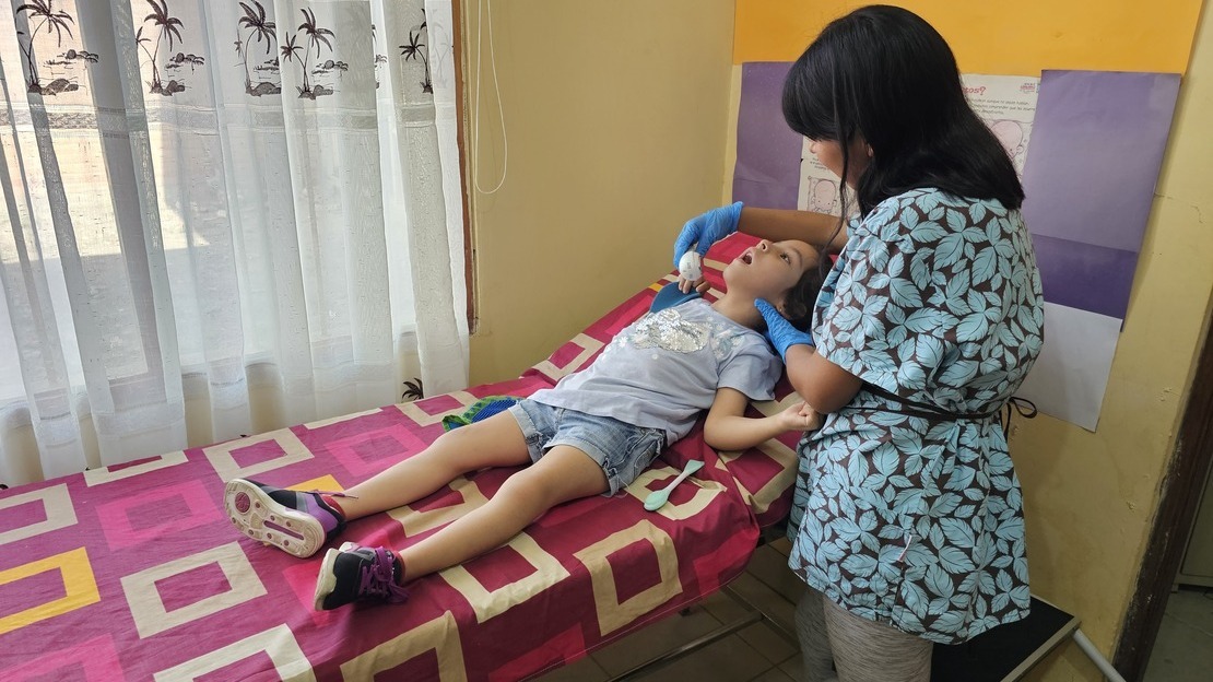 Tamara est allongée sur un lit de clinique. A côté d'elle, se tient Laura Giovana qui tient à la main un masseur facial qu'elle passe délicatement sur la joue de la fillette.