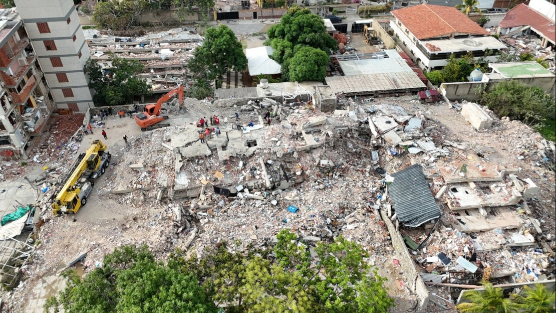 A drone view of a group of buildings that have been completely destroyed. Service vehicles and people can be seen amidst the rubble.