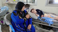 Un petit garçon dont les jambes sont recouvertes de bandage est allongé sur un lit d'hôpital. Sa grand-mère est assise à ses côtés. 