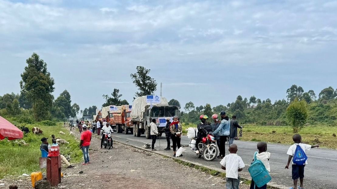 Une file de camion affichant les logo de HI et de l'UE avancent sur une route. Sur les bas-côtés, de nombreuses personnes en moto ou à pieds transportant des paquet ou regardant passer les camions.