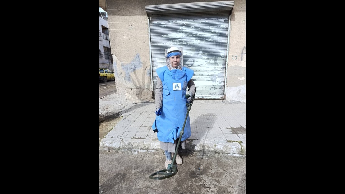 Une femme se tient debout sur un trottoir devant un rideau métallique fermée. Elle porte un gilet de protection épais de couleur bleu et une visière transparente. Elle tient un détecteur de métaux orienté vers le sol. Le bâtiment derrière est usé, avec des murs partiellement endommagés. Une voiture jaune est visible sur la gauche en arrière plan dans la rue.