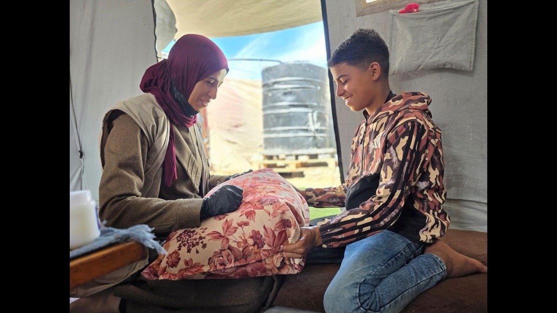 À l’intérieur d’un abri en toile, deux personnes sont assises face à face sur des coussins. Une personne adulte porte des vêtements superposés et un foulard couvrant les cheveux, et tient un coussin à motif floral posé sur les genoux. En face, une personne plus jeune, agenouillée sur un tapis, porte un sweat à capuche à motifs et un pantalon en jean, et maintient également le coussin avec les mains. À l’arrière-plan, on voit une ouverture laissant apparaître un ciel bleu et des textiles accrochés. La lumière naturelle éclaire la scène depuis l’ouverture.