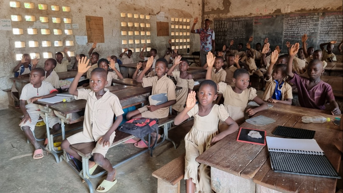 Intérieur d'une salle de classe. Des enfants en uniformes beige âgés de 8-10 ans sont assis autour de pupitres en bois et lèvent la main.