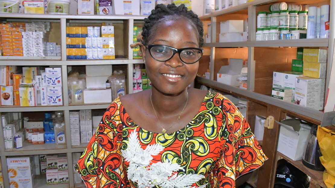 Portrait d'une jeune femme qui pose dans une pharmacie, avec des rayonnages remplis de boîtes de médicaments. La jeune femme porte des lunettes et sourit à la caméra.