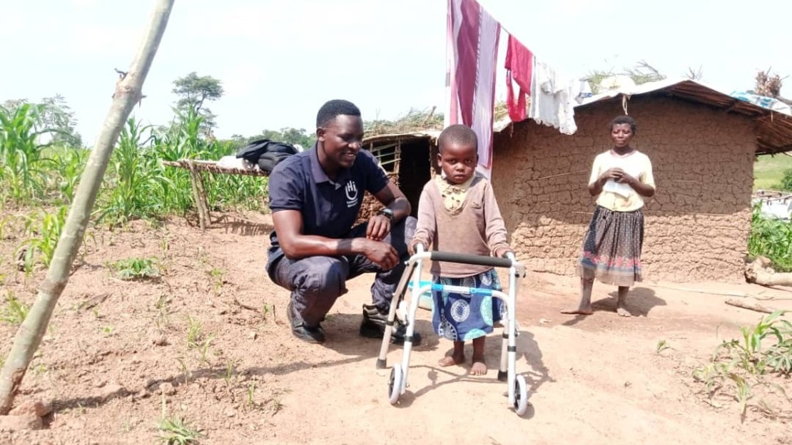 In front of a brick house, a little girl stands leaning on a walking frame. Kneeling beside her is a smiling man. A woman stands in the background.