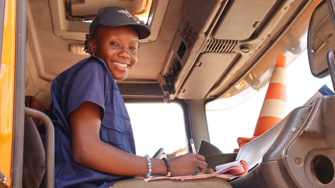 Une jeune femme noire est assise dans une cabine de camion. Elle tient un stylo et des papiers et regarde la caméra en contrebas en souriant.