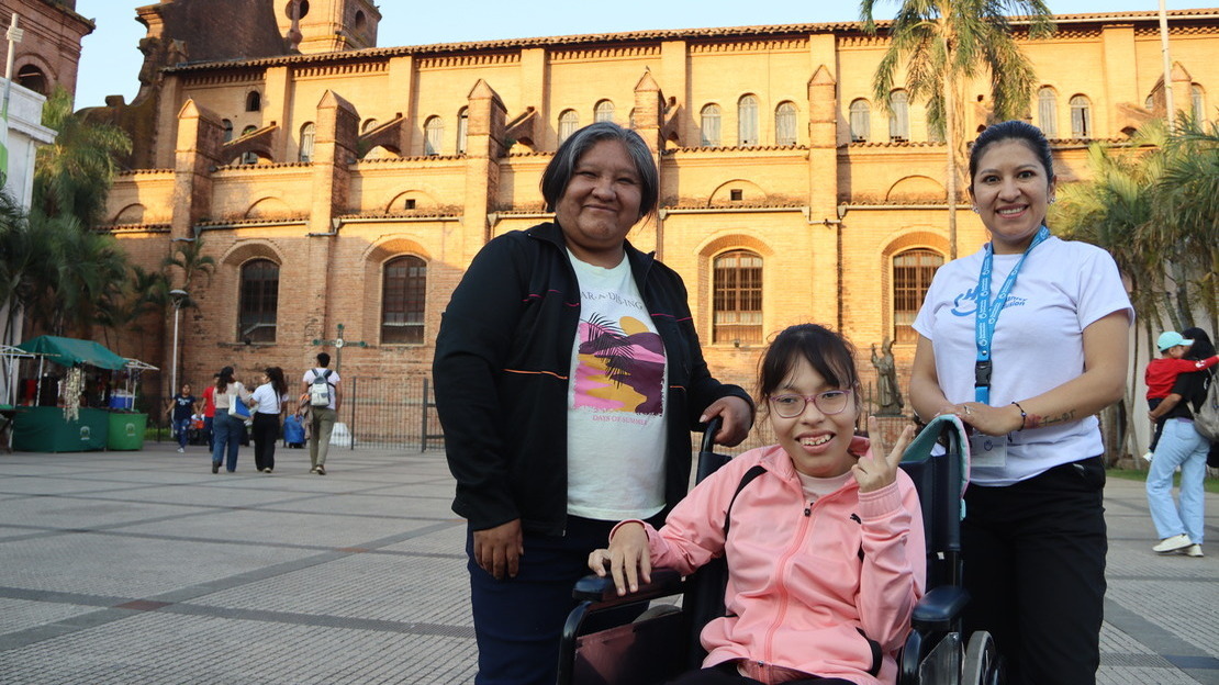 Trois femmes se tiennent sur une place, devant une cathédrale illuminée par le soleil. Deux d'entre elles sont debout et entourent la troisième, assises sur un fauteuil roulant. Elles sourient à la caméra.