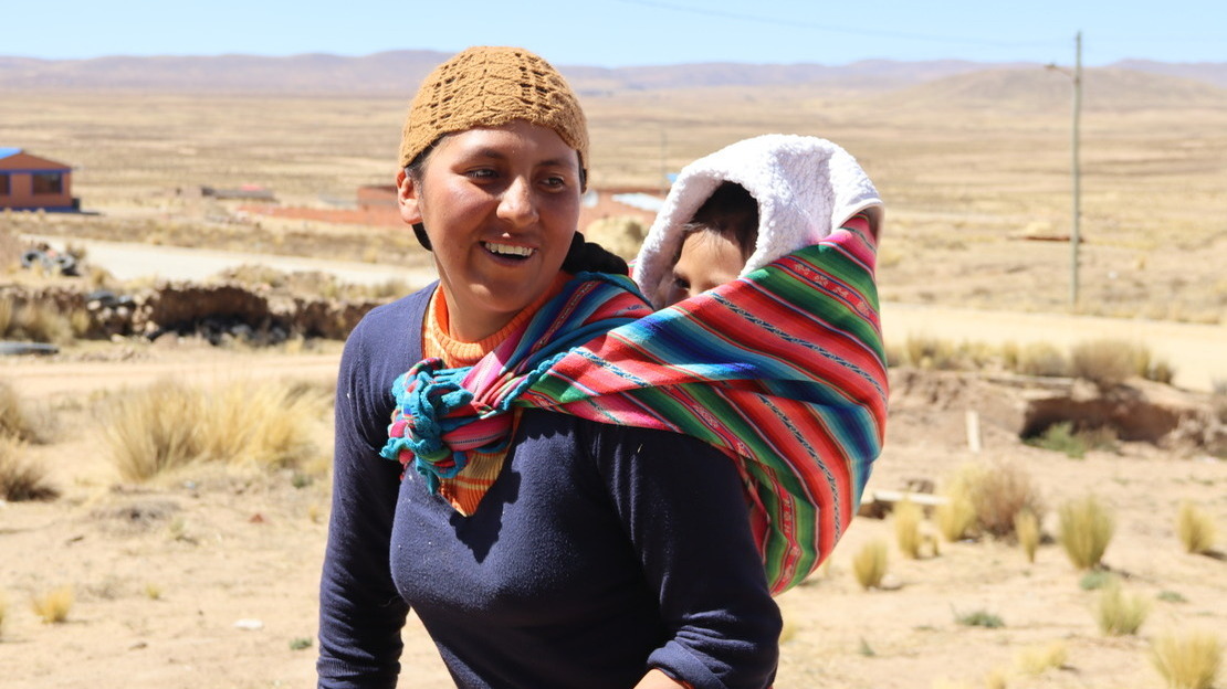 Une femme souriante porte son fils dans son dos à l'aide d'un tissu traditionnel (aguayo). En arrière-plan, les plaines sèches et poussiéreuses de l'Altiplano.