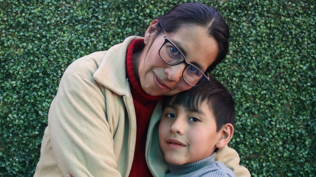 Maria and Hector are hugging in front of a vegetal wall 