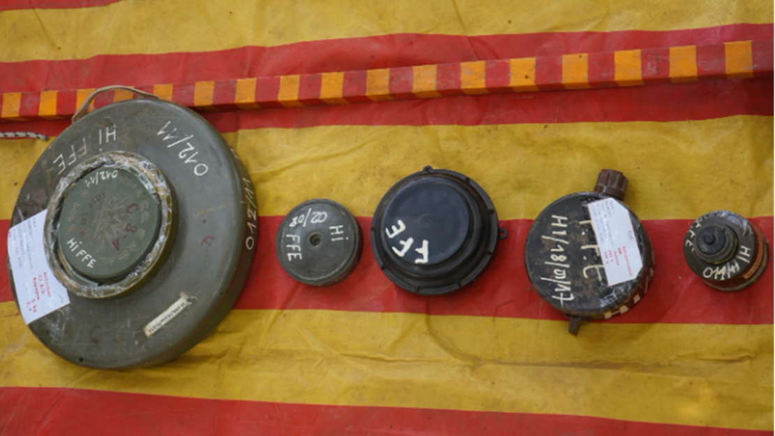 5 objets métalliques sont disposés en ligne sur une bâche jaune avec de larges bandes rouges. Les objets ont des formes circulaires ou cylindriques et présentent des surfaces usées et oxydées. Ces objets sont de tailles différentes et porte des inscriptions blanches peintes à la main. L’ensemble des objets est présenté de manière ordonnée sur la bâche, dans un environnement extérieur ou temporaire, avec un éclairage naturel.