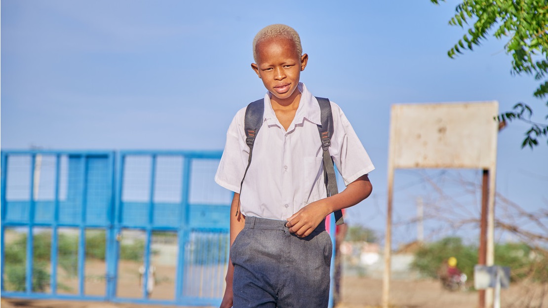 Portrait d'un adolescent en uniforme scolaire qui entre dans la cour d'une école en souriant. Derrière lui, on devine un portail en fer forgé bleu, un arbre et un grand ciel bleu sans nuage.