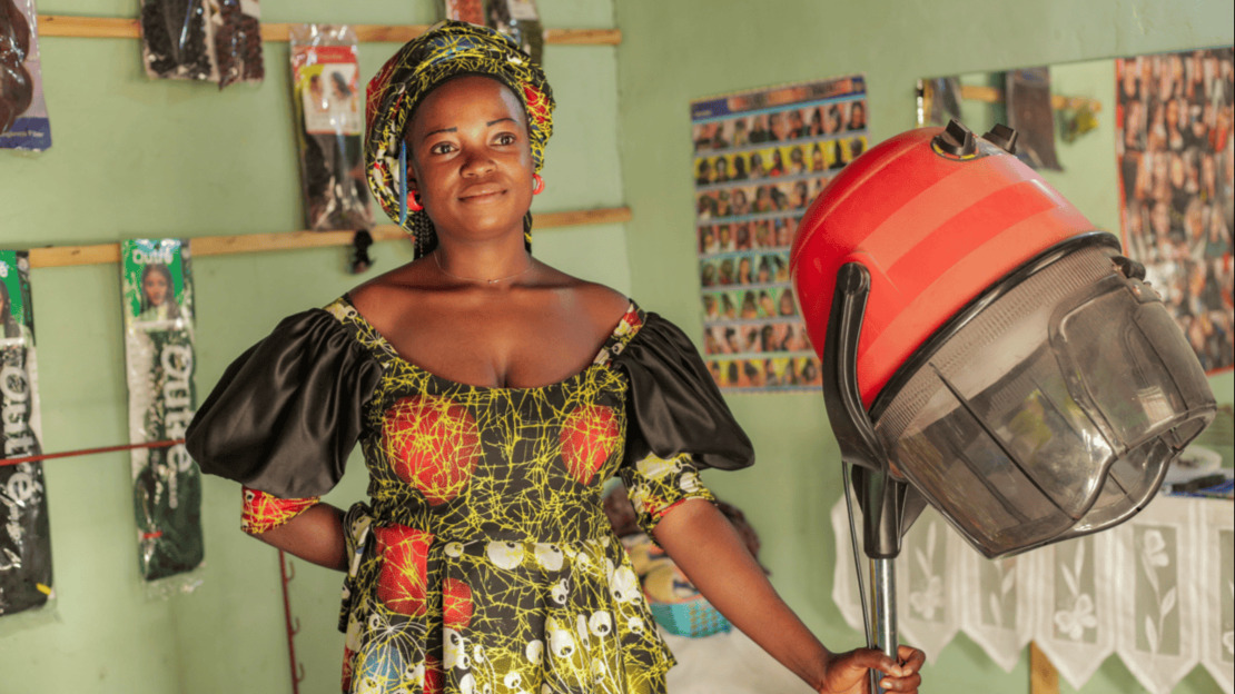 A smiling young woman is standing in a room whose walls are covered with posters of hairstyles and locks of hair. She is holding a hairdryer.