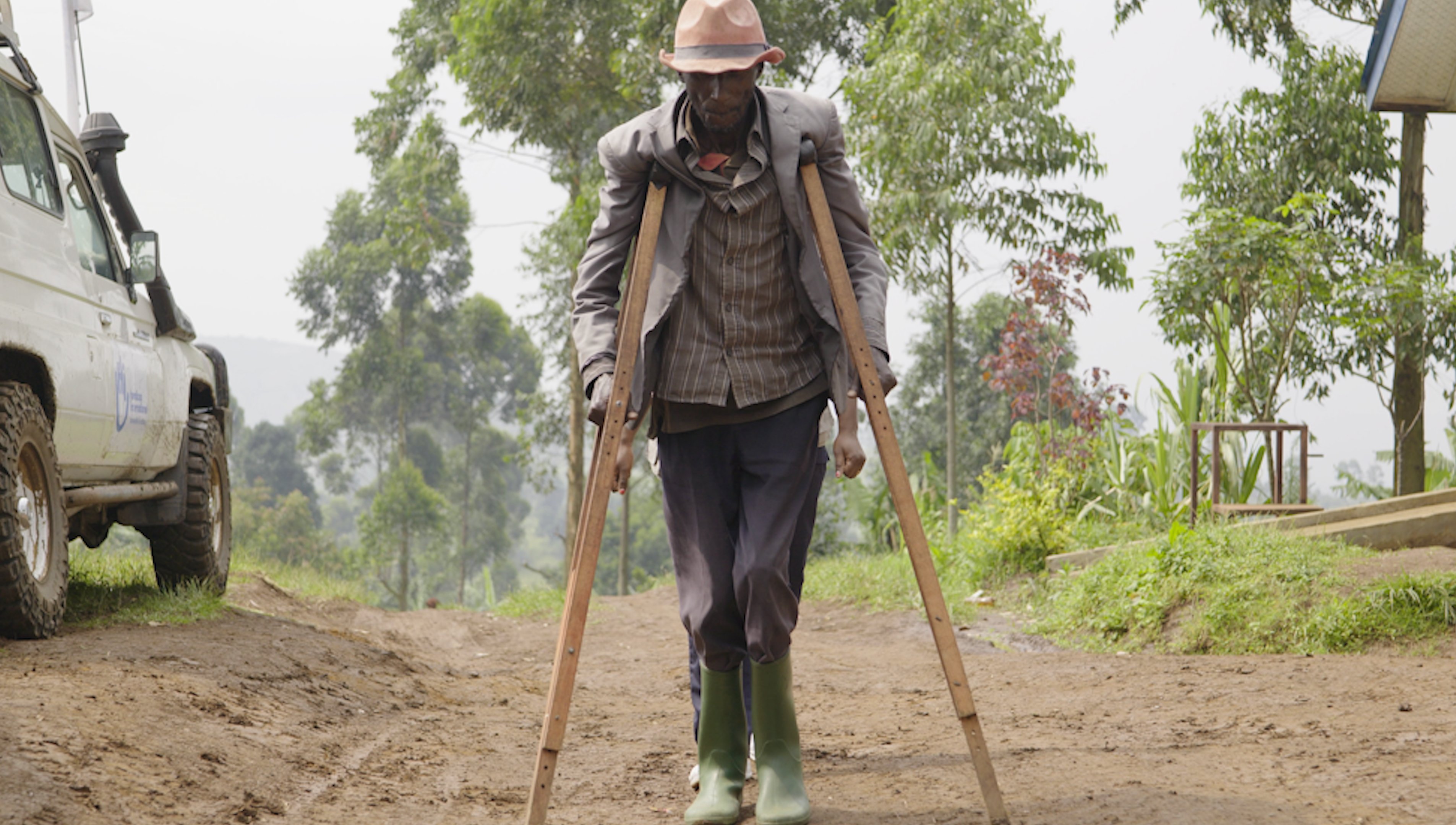Une personne avance sur un chemin en terre en s’appuyant sur deux longues béquilles en bois, placées de chaque côté du corps. La personne porte une veste, une chemise en rayure, un pantalon et des bottes en caoutchouc vertes, ainsi qu’un chapeau. Le chemin traverse un environnement bordé d’arbres et de plantes. Sur le côté gauche de l’image, un véhicule blanc est partiellement visible, stationné le long du chemin. En arrière-plan, une autre personne est visible derrière la personne.