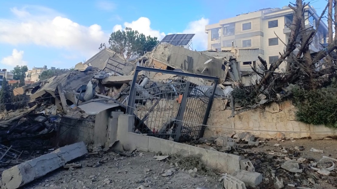 L’image montre un bâtiment effondré, avec des blocs de béton brisés, une grille métallique tordue et des arbres endommagés au premier plan. En arrière plan, on voit d’autres immeubles intacts sous un ciel nuageux. 