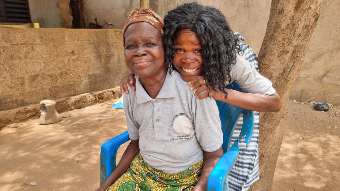 Une jeune femme enlace par derrière une femme plus âgée assise sur une chaise, dans une cour en pisé. Elle sourient toutes les deux en regardant la caméra.