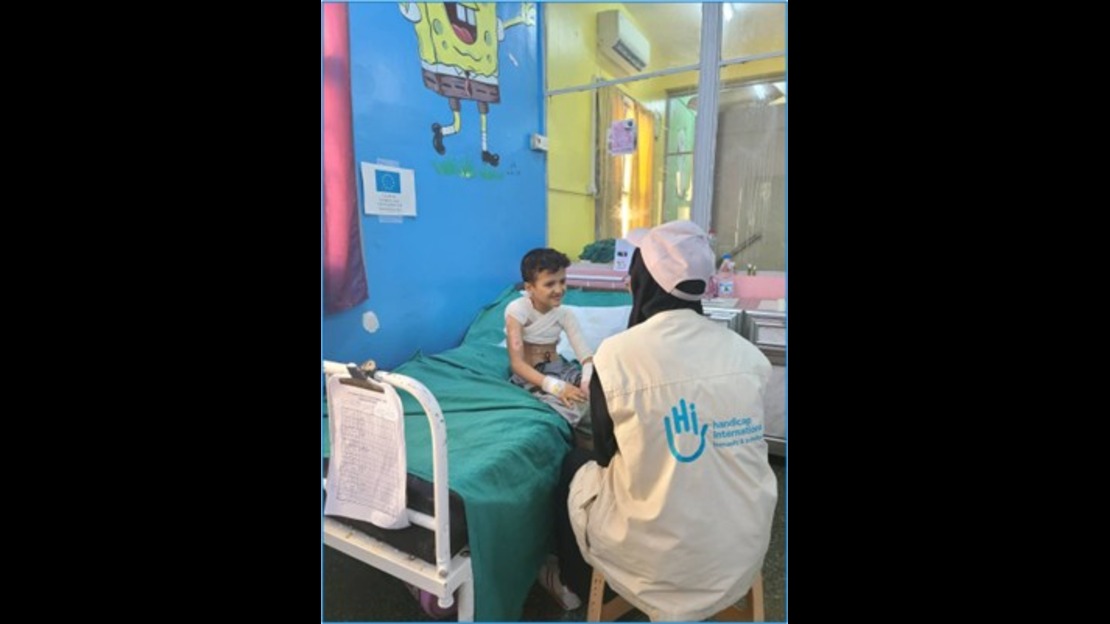 Un enfant est assis sur un lit médical recouvert d’un drap vert dans une pièce d’hôpital colorée, face à une personne portant un gilet identifiable au logo. La chambre comprend du mobilier médical, des documents posés sur le lit et une décoration murale illustrée.
