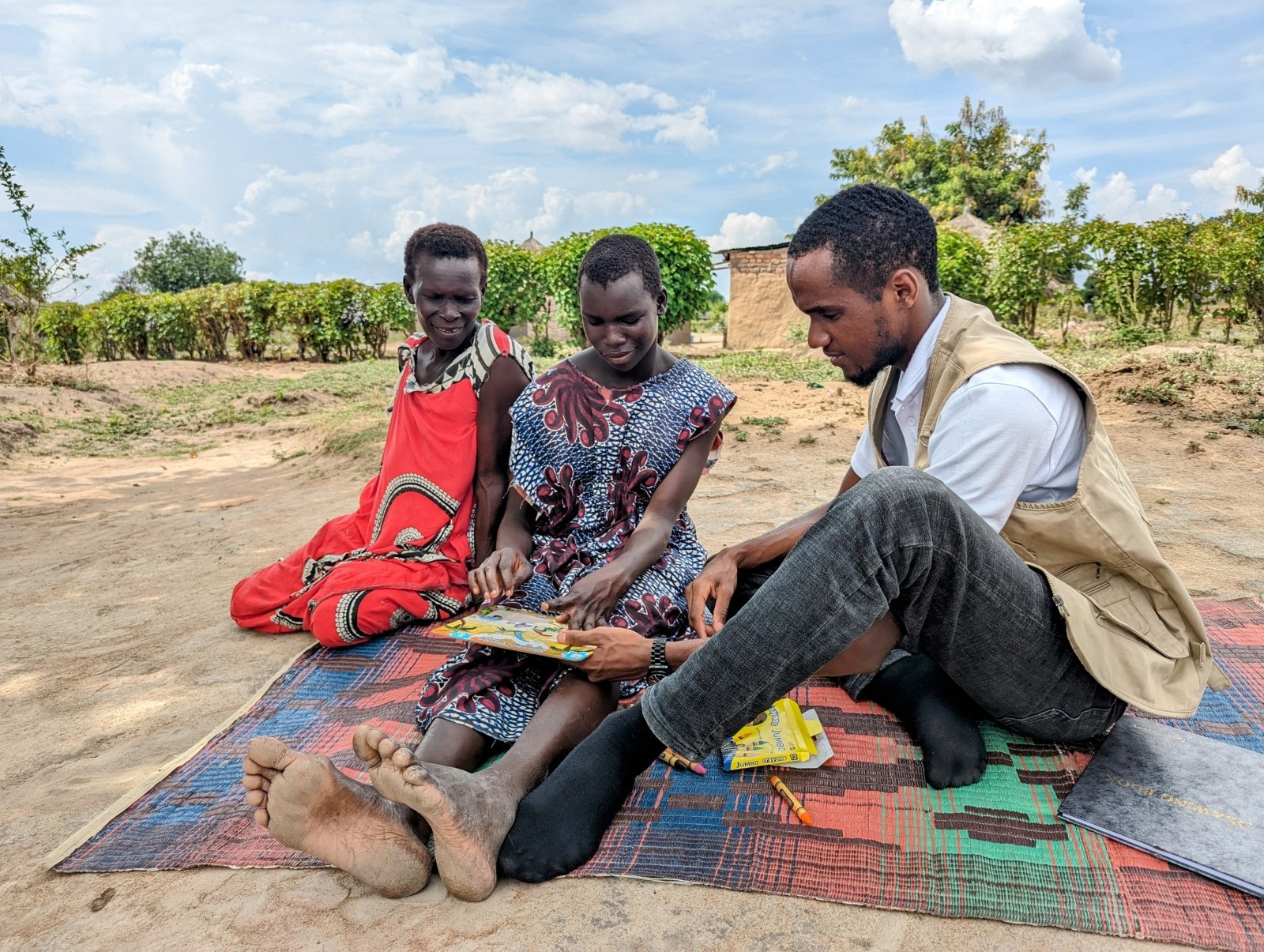Santina faisant un puzzle, accompagnée de sa mère et d'un ergothérapeute de chez HI. Ils sont assis tous les trois sur un tapis, à l'extérieur. Santina est au centre de l'image, et sa mère et l'égothérapeute, qui l'entourent,  la regardent en faire le puzzle en souriant. 