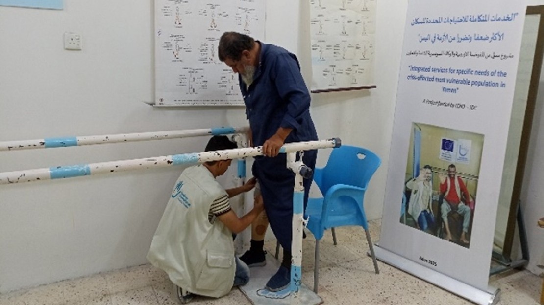 Une personne se tient debout entre des barres parallèles de rééducation dans une salle. Une autre personne ajuste une prothèse de jambe au niveau de son membre inférieur. La salle comprend des affiches techniques, une chaise en plastique bleue et un panneau d’information sur l’inclusion et la réadaptation.