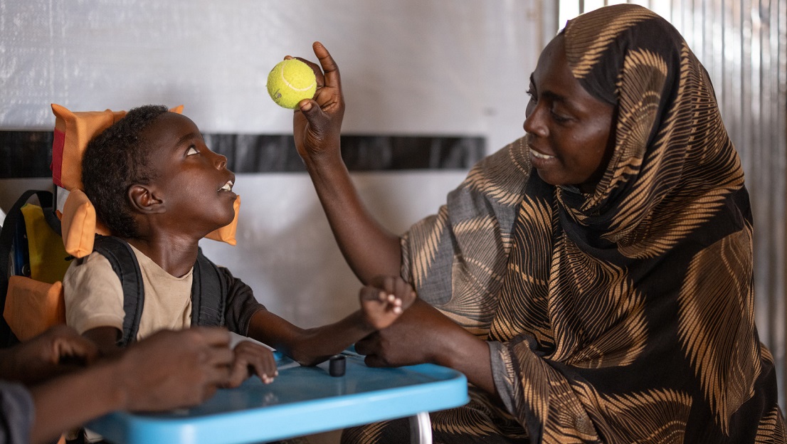 La photo est en intérieur, dans une salle de réadaptation d'HI. Omran, à gauche, est dans un fauteuil adapté et regarde la maman qui lui montre une balle de tennis. Djimilla, sa maman, regarde Omran en souriant. 