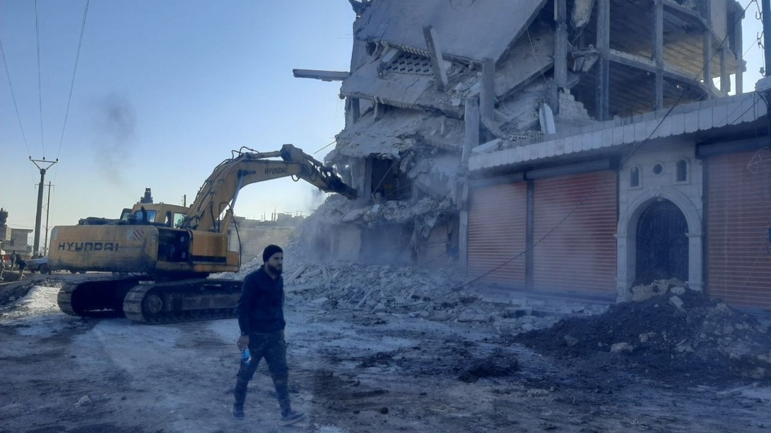 Un homme marche devant une pelleteuse qui creuse un immeuble effondré