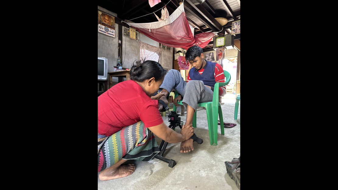 Dans une pièce d’habitation au sol en ciment, une personne accroupie ajuste un dispositif placé au niveau du pied et de la cheville d’une autre personne assise sur une chaise verte en plastique. La personne assise a les jambes posées devant elle et observe l’intervention. L’arrière plan montre des murs en béton décorés de calendriers et d’images, un téléviseur posé sur un meuble, ainsi que plusieurs tissus suspendus au plafond.