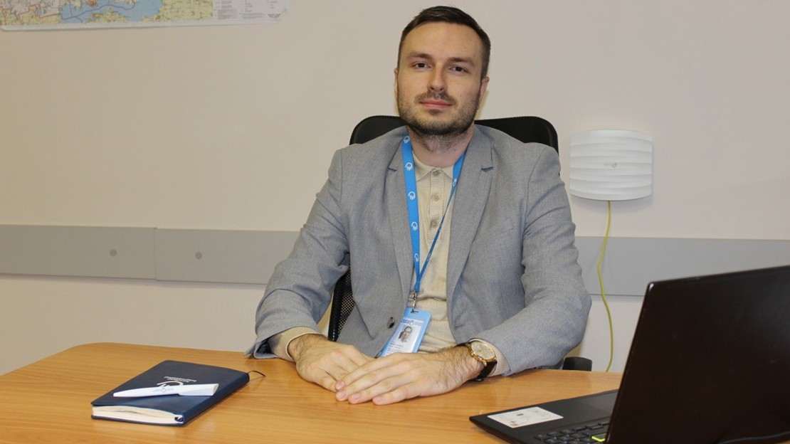 Une personne est assise à un bureau dans un environnement de travail. Elle porte une veste claire et un badge avec un cordon bleu avec un logo visible. Ses mains sont posées sur le bureau, sur lequel se trouvent un carnet et un ordinateur portable ouvert. En arrière-plan, un mur clair avec une carte accrochée et un appareil mural.