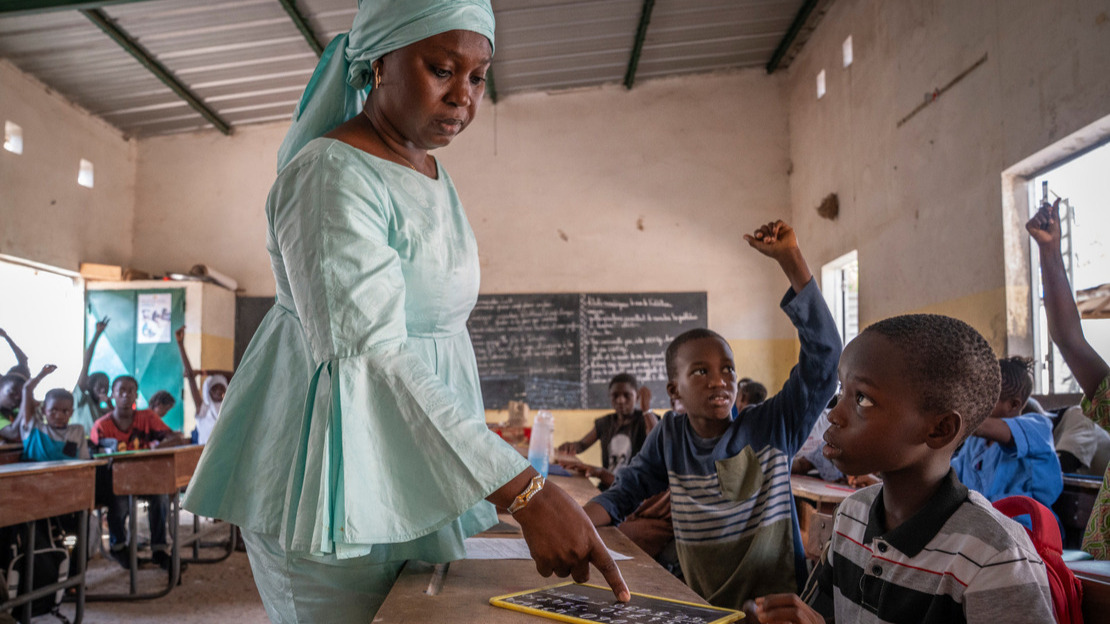 Dans une salle de classe, une femme montre quelque chose sur l'ardoise d'un garçon assis au premier plan.