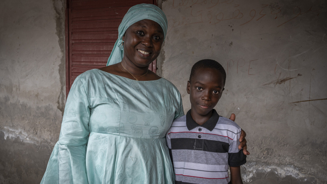 Portrait en gros plans d'une femme qui tient par l'épaule un jeune garçon. Tous les deux regardent la caméra et sourient.