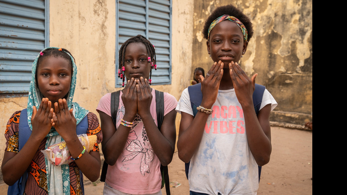 Trois jeunes filles se tiennent debout côte à côte et portent chacune leurs deux mains au niveau du bas de leur visage, paumes ouvertes tournées vers elles.