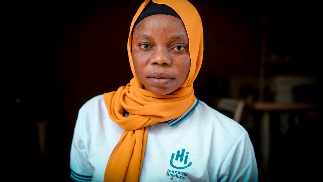 Portrait de Saly : elle porte un foulard orange et un t-shirt avec le logo de HI. En arrière-plan, on devine son atelier de couture.