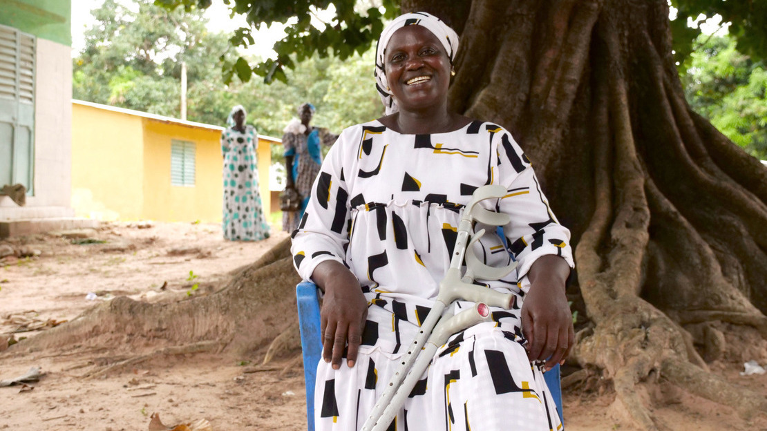 Une femme est assise sur une chaise en plastique bleu, posée devant un grand arbre fromager. Elle est habillée d'une robe blanche aux motifs noir et jaune et sourit à la caméra. Des béquilles sont appuyées sur ses jambes.