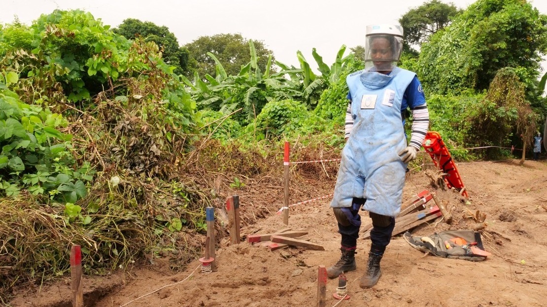 Une femme en tenue de démineuse se tient devant un couloir de déminage marqué par des piquets bleu et rouge, sur un chemin de terre. Derrière, de la végétation luxuriante.