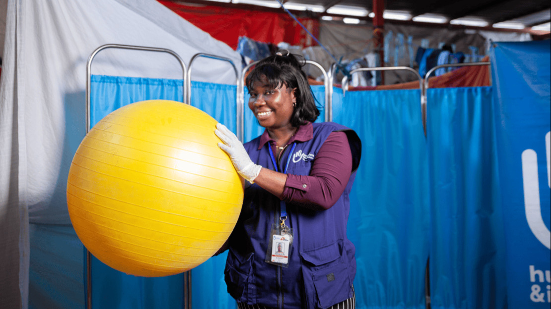 Portrait of a smiling woman wearing a latex glove and a HI jacket, holding a large yellow physiotherapy ball.