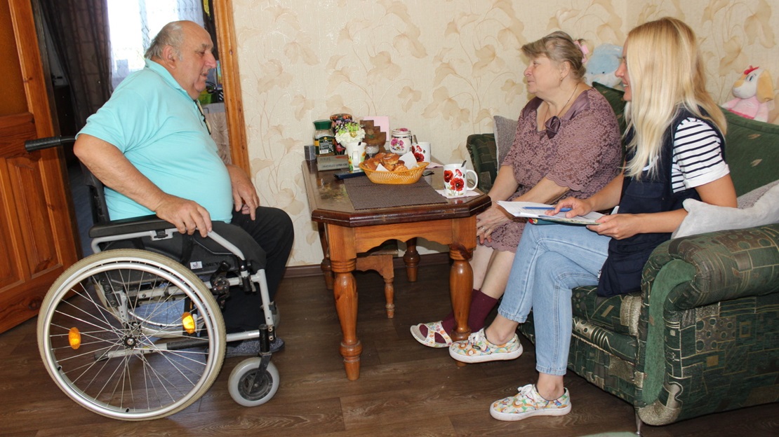 Trois personnes sont autour d'une table basse, un homme est en fauteuil roulant. Ils discutent.