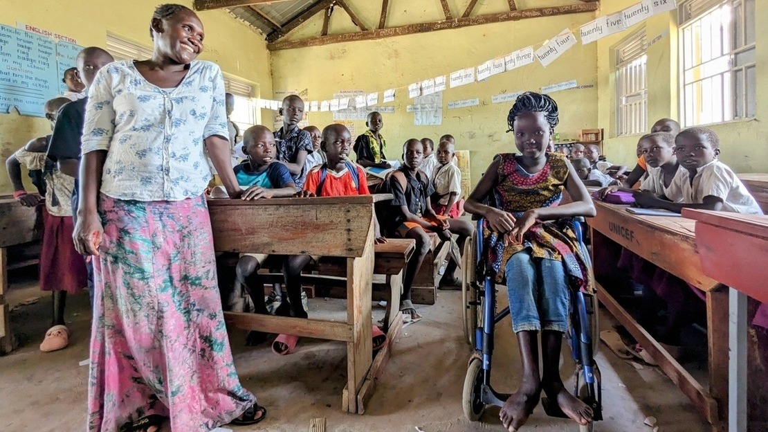 Vue d'une salle de classe avec des élèves assis à des pupitres. Au premier plan, une femme adulte qui sourit en regardant à sa gauche une jeune fille assise sur un fauteuil roulant.