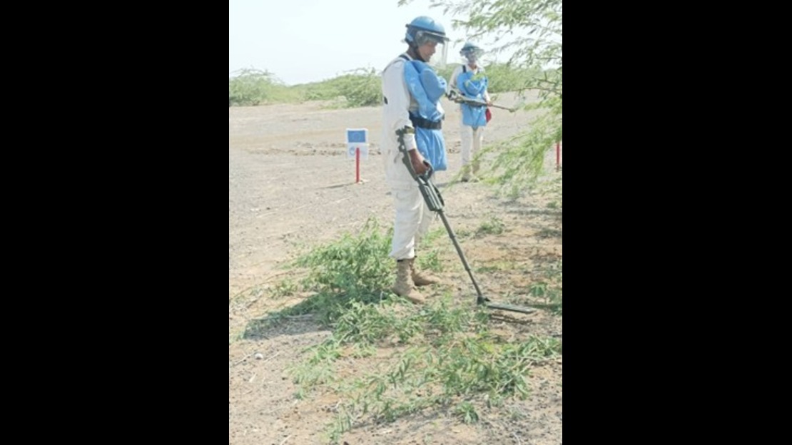Two people wearing protective helmets, vests, and metal detectors are inspecting an open, dry outdoor area with low vegetation. Marker stakes are visible, indicating a technical work zone.