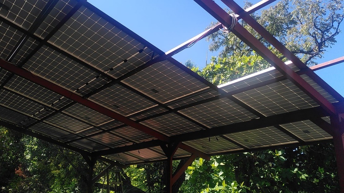 Vue en contre-plongée d'une structure métallique presque entièrement recouverte de panneaux solaire. À travers les emplacements encore vides, on voit des arbres et du ciel bleu.