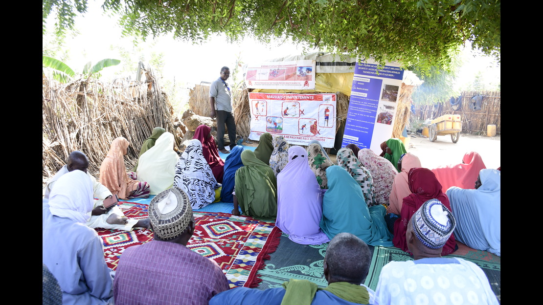 Un groupe de personnes est assis au sol sur des tapis et nattes aux motifs colorés, disposés à l’ombre d’un grand arbre dans un environnement rural. Une personne se tient debout devant le groupe et présente des informations à l’aide de plusieurs affiches et panneaux fixés à une structure faite de matériaux simples. Les supports visuels comprennent des illustrations, des pictogrammes et des blocs de texte. À l’arrière-plan, une clôture en branches entoure l’espace, et un chemin ainsi qu’une charrette sont visibles. 
