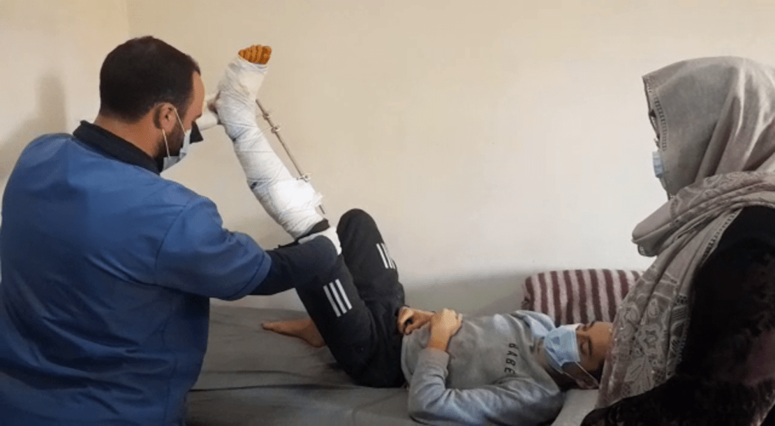 Un adolescent est allongé sur son lit d'hôpital, sa jambe est cassée et le kinésithérapeuthe lui fait faire des exercices de rééducation avec sa mère pour le soutenir à coté.